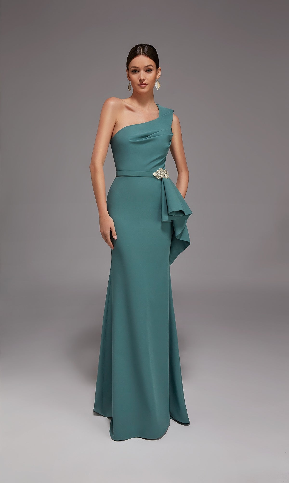 A woman stands in a studio wearing the Alyce 27812 long formal dress—a dark sage, one-shoulder, floor-length gown with a side drape and crystal beaded accent brooch at the waist.